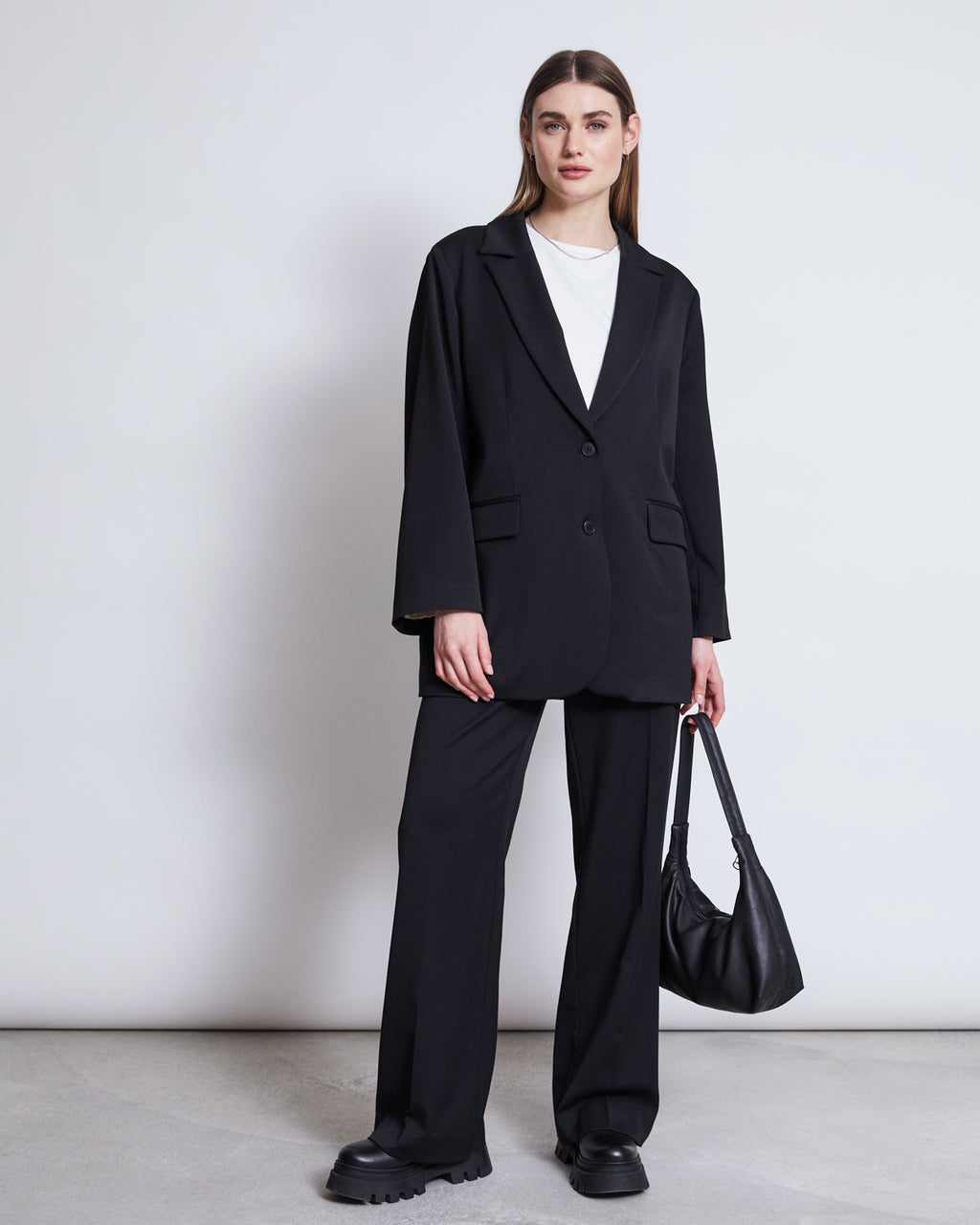 A woman with straight brown hair tucked behind her ears stands against a plain white wall wearing jan-n-junes BLAZER DURANCE BLACK, matching wide-leg trousers, a white T-shirt, black chunky shoes, and holding a black handbag.