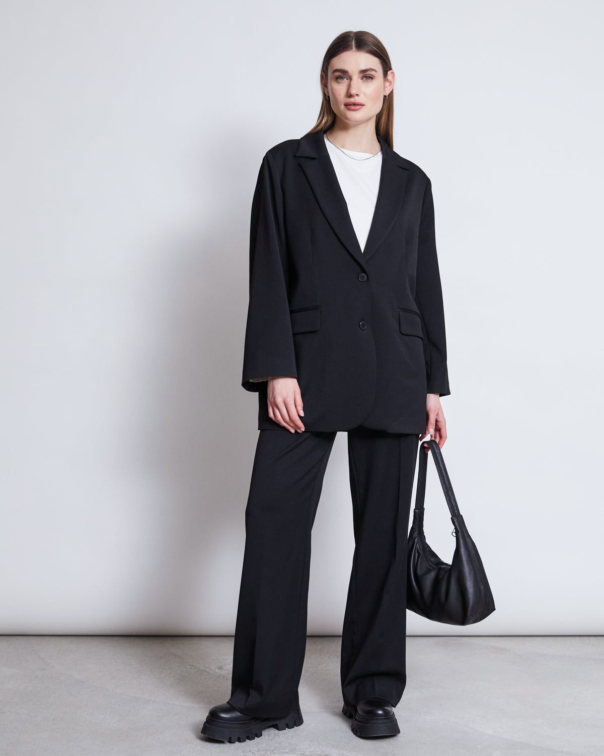 A woman with straight brown hair tucked behind her ears stands against a plain white wall wearing jan-n-junes BLAZER DURANCE BLACK, matching wide-leg trousers, a white T-shirt, black chunky shoes, and holding a black handbag.