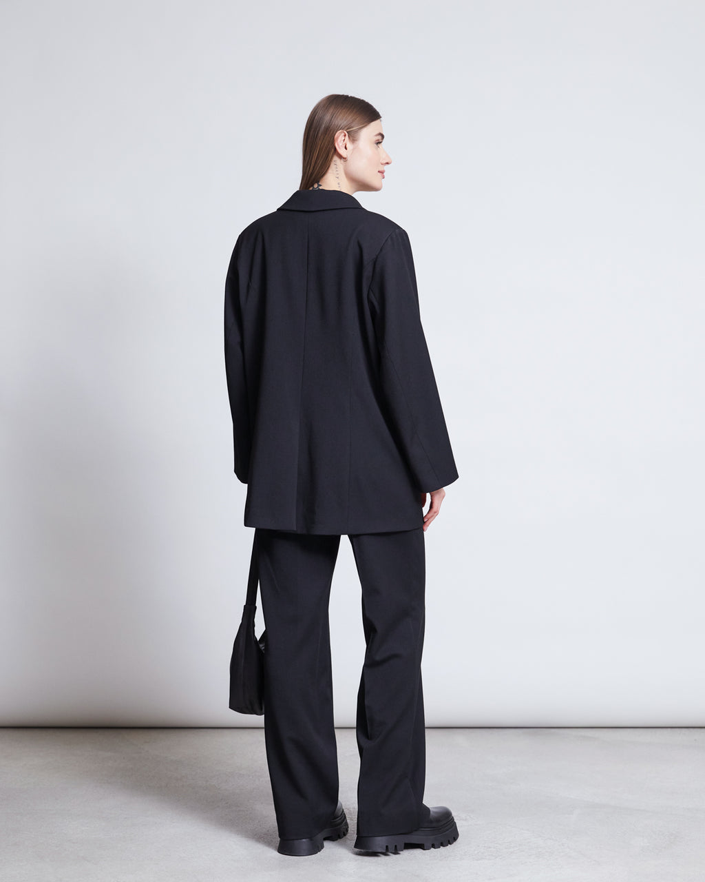 A woman with straight brown hair faces away, wearing jan-n-junes BLAZER DURANCE BLACK—a recycled polyester oversized blazer—paired with matching pants, chunky black shoes, and a small black bag against a plain light background.
