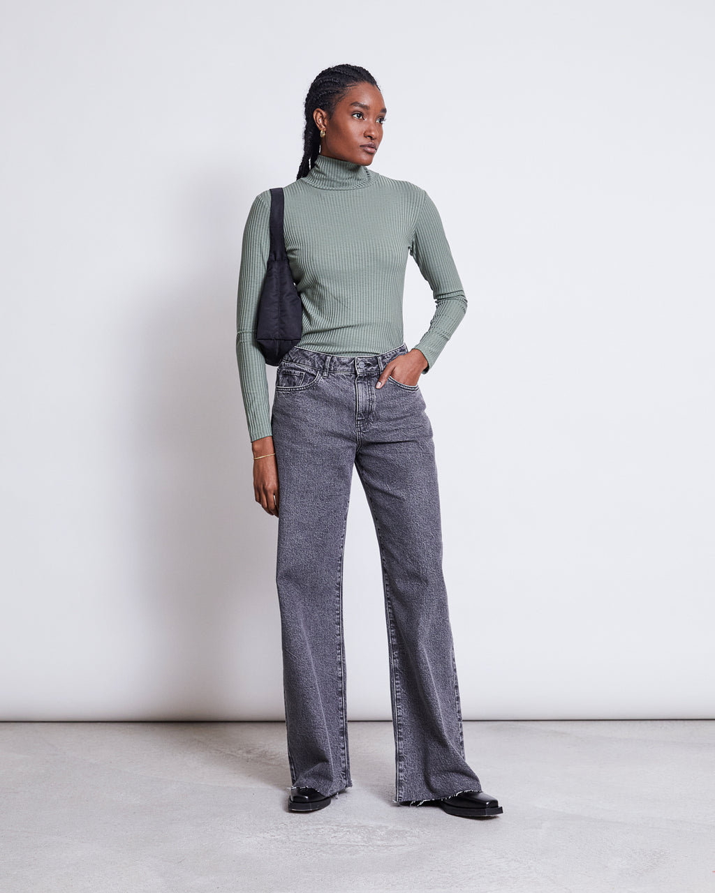 A person stands against a white background wearing the jan-n-june LYOCELL RIB TURTLENECK MIO SEA SPRAY, paired with gray high-waisted wide-leg jeans, black shoes, and a black shoulder bag, one hand in their pocket.