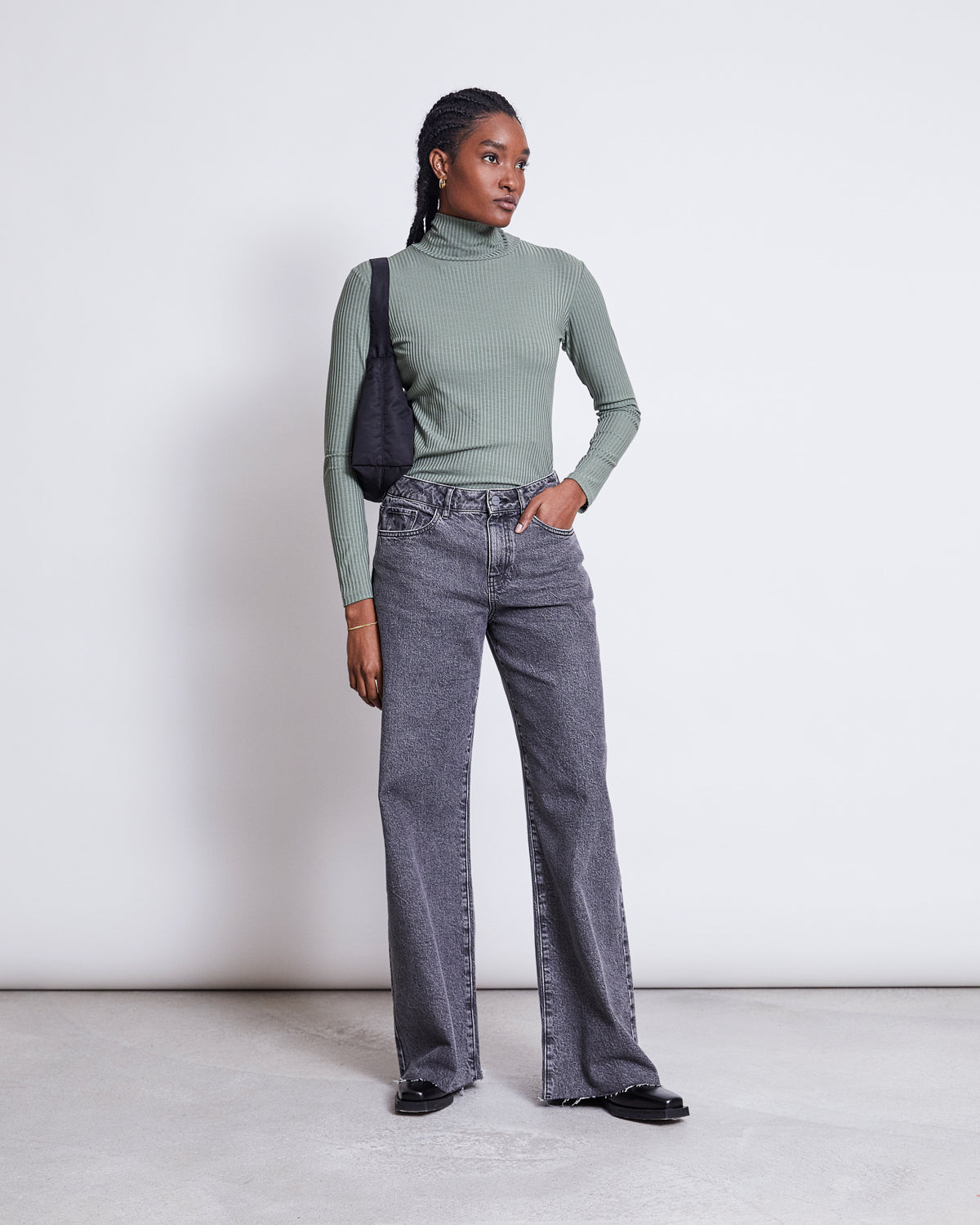 A person stands against a white background wearing the jan-n-june LYOCELL RIB TURTLENECK MIO SEA SPRAY, paired with gray high-waisted wide-leg jeans, black shoes, and a black shoulder bag, one hand in their pocket.