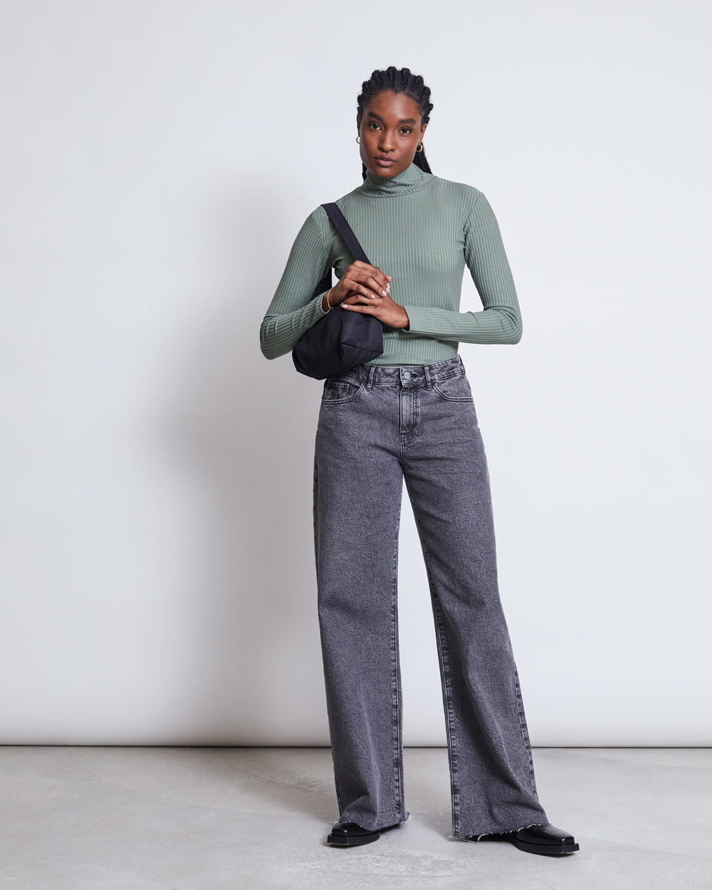 A woman with braided hair poses confidently against a plain light background in the jan-n-june LYOCELL RIB TURTLENECK MIO SEA SPRAY, gray wide-leg jeans, black shoes, and a black shoulder bag.