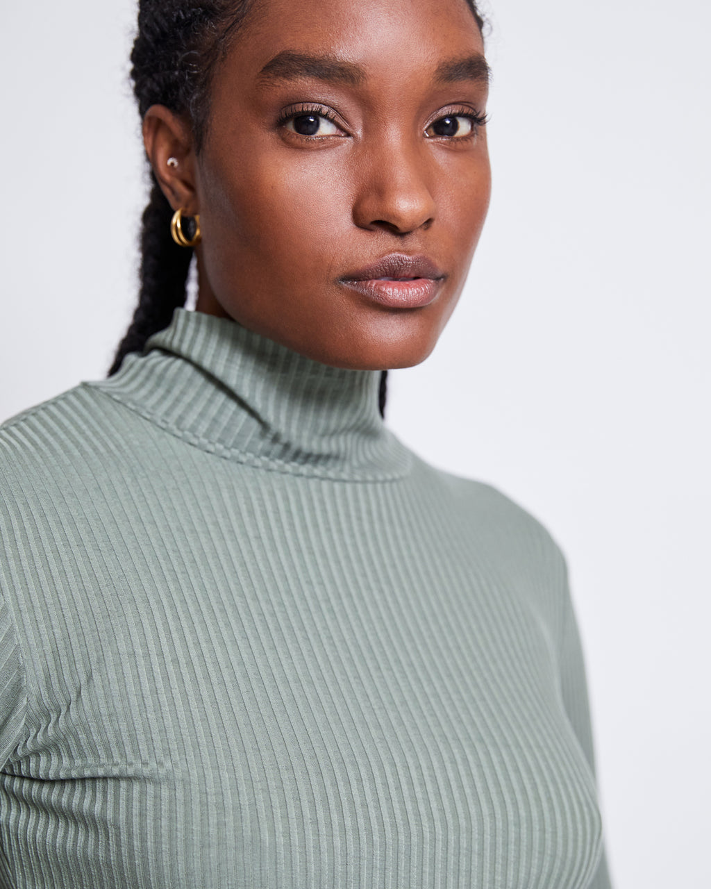 A woman with braided hair and gold hoops wears the jan-n-june LYOCELL RIB TURTLENECK MIO SEA SPRAY, confidently facing the camera against a plain light background.