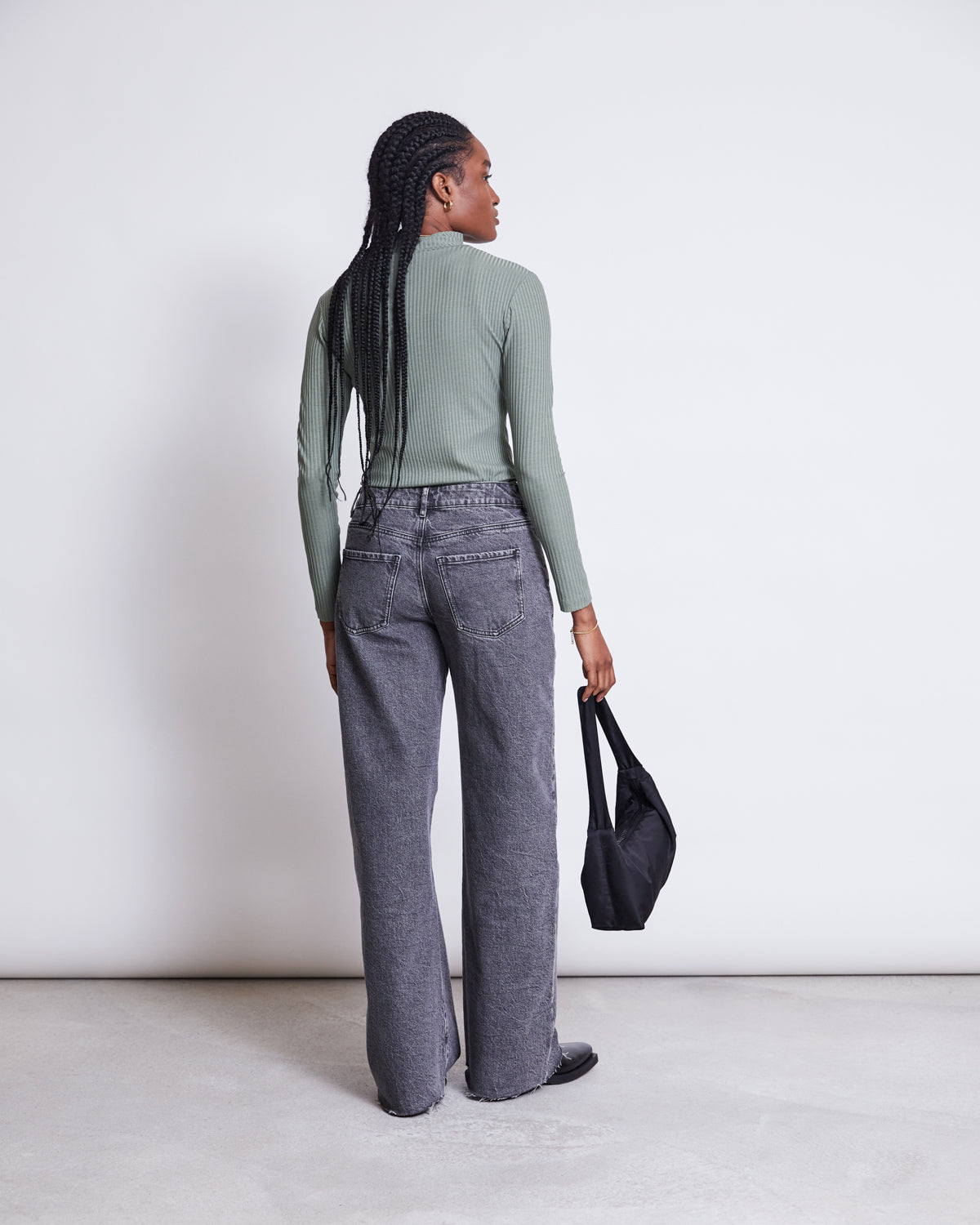 A person with long braided hair wears the jan-n-june LYOCELL RIB TURTLENECK MIO SEA SPRAY green top, loose gray jeans, and black shoes, facing away and holding a black handbag against a plain white background.
