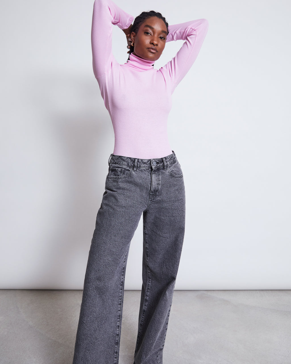 A person stands confidently against a plain background in jan-n-junes WIDE LEG JEANS SUNNY LIGHT GREY, styled with a light pink turtleneck, hands behind their head for an effortless look and comfortable wear.