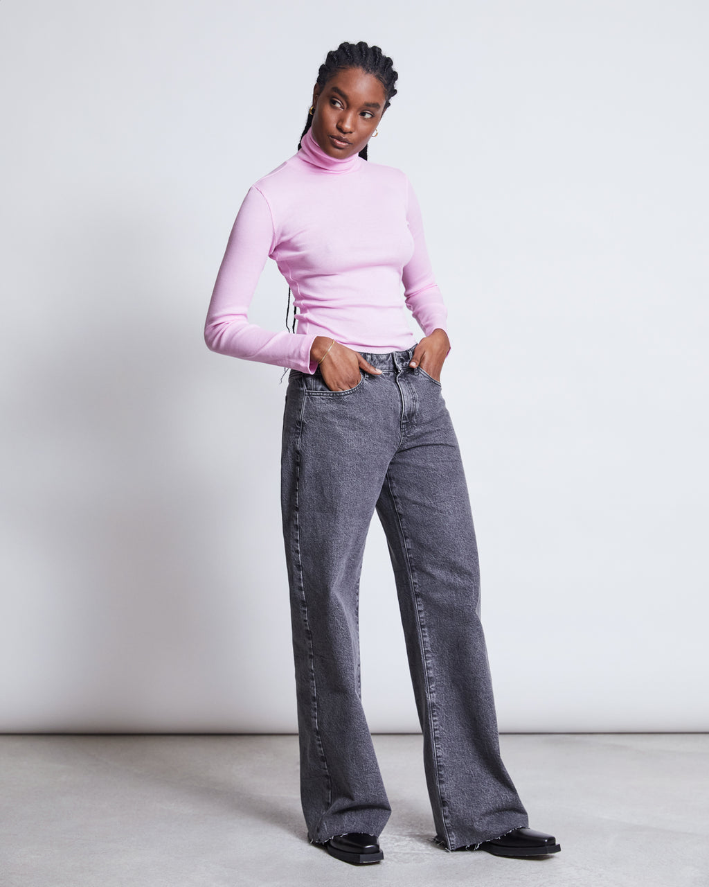 A person with braided hair wears jan-n-junes WIDE LEG JEANS SUNNY LIGHT GREY made from organic cotton and a light pink turtleneck, standing hands-in-pockets against a plain background for high comfort.