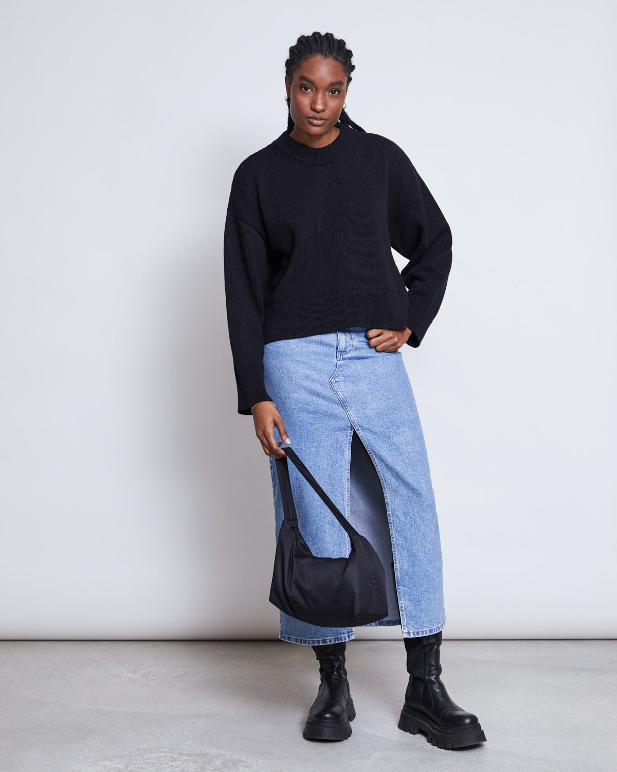 A person stands against a plain background wearing the jan-n-june JUMPER YAGMUR BLACK GOTS, a light blue denim skirt with a front slit, black boots, and holding a small black shoulder bag.