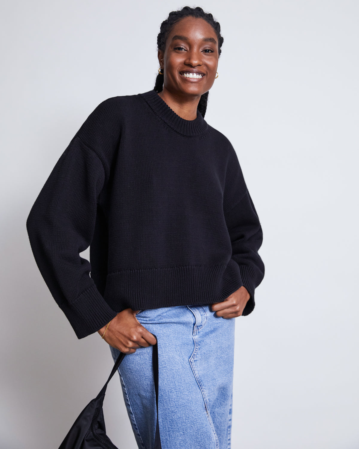 A woman with braided hair smiles in front of a plain light background, wearing the jan-n-june JUMPER YAGMUR BLACK GOTS and light blue jeans. One hand is in her pocket while the other holds a black strap.