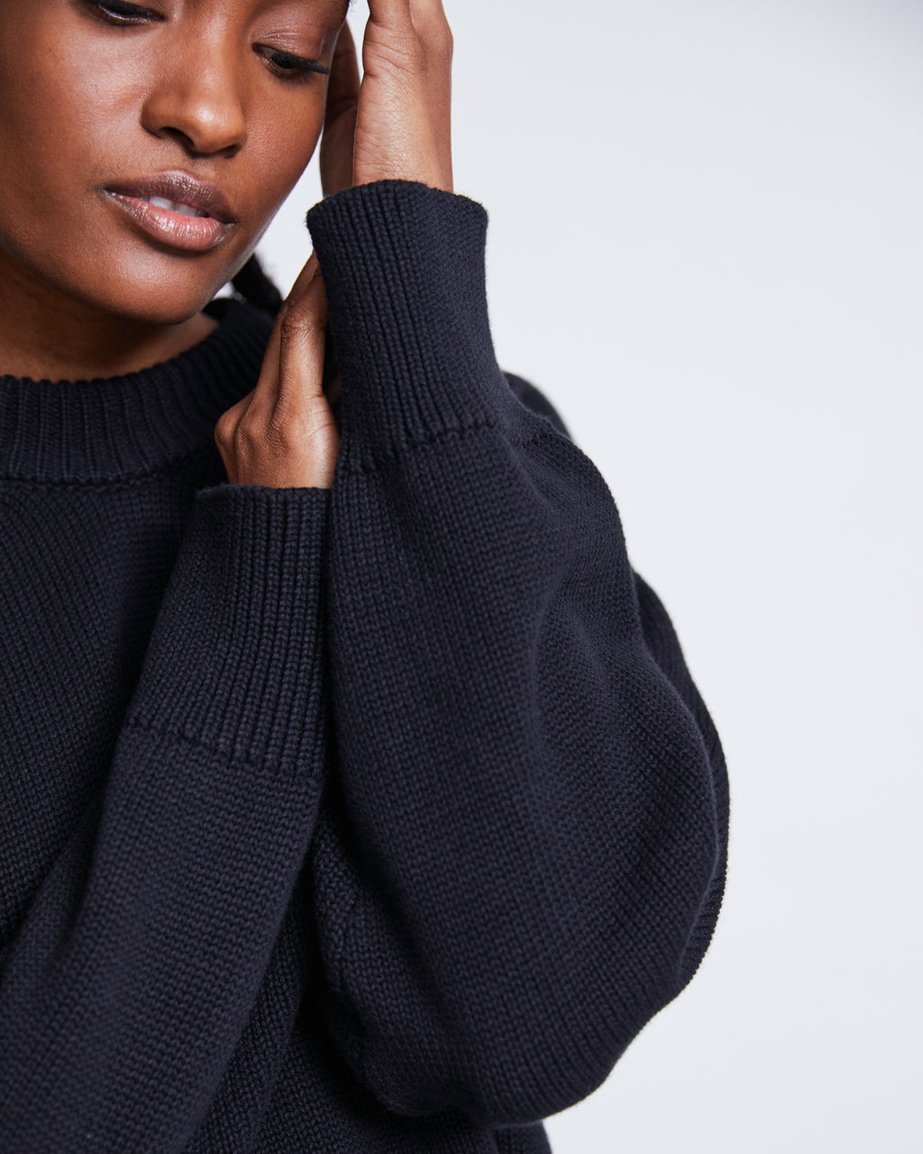 Wearing the jan-n-june JUMPER YAGMUR BLACK GOTS, a person gently touches their face with one hand, eyes partly closed, and only half of their face visible in this close-up image.