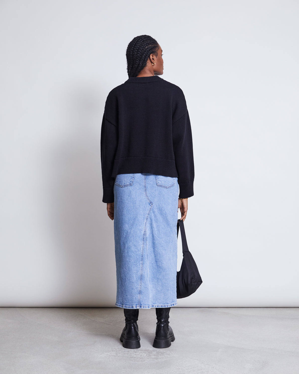 A person with braided hair faces away, wearing the jan-n-june JUMPER YAGMUR BLACK GOTS, a long light blue denim skirt, black boots, and holding a black bag by their side against a plain light background.