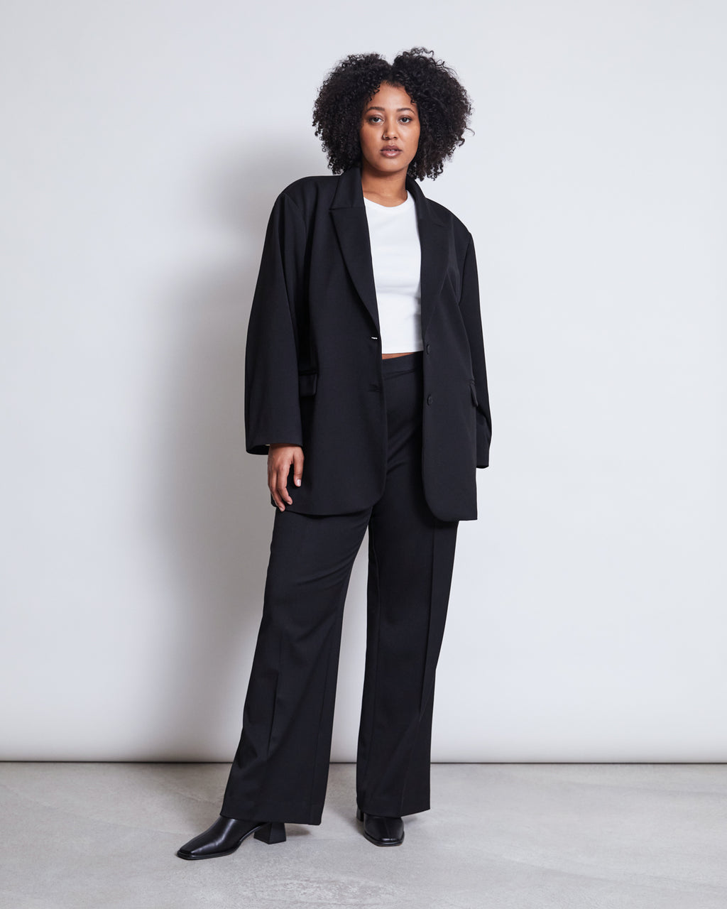 A woman with curly hair stands against a plain background, wearing the jan-n-june BLAZER DURANCE BLACK—an oversized blazer crafted from recycled polyester—paired with matching black trousers, a white crop top, and black heeled boots.