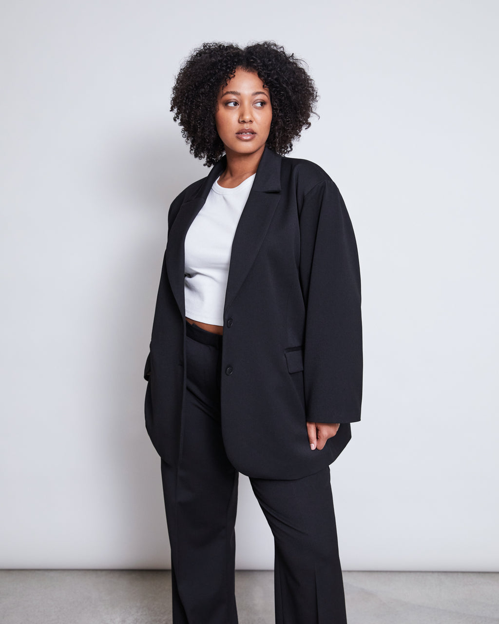 A woman with curly hair wears the jan-n-june BLAZER DURANCE BLACK, an oversized blazer made from recycled polyester, paired with matching pants and a white crop top. She stands against a plain light background, looking to the side.