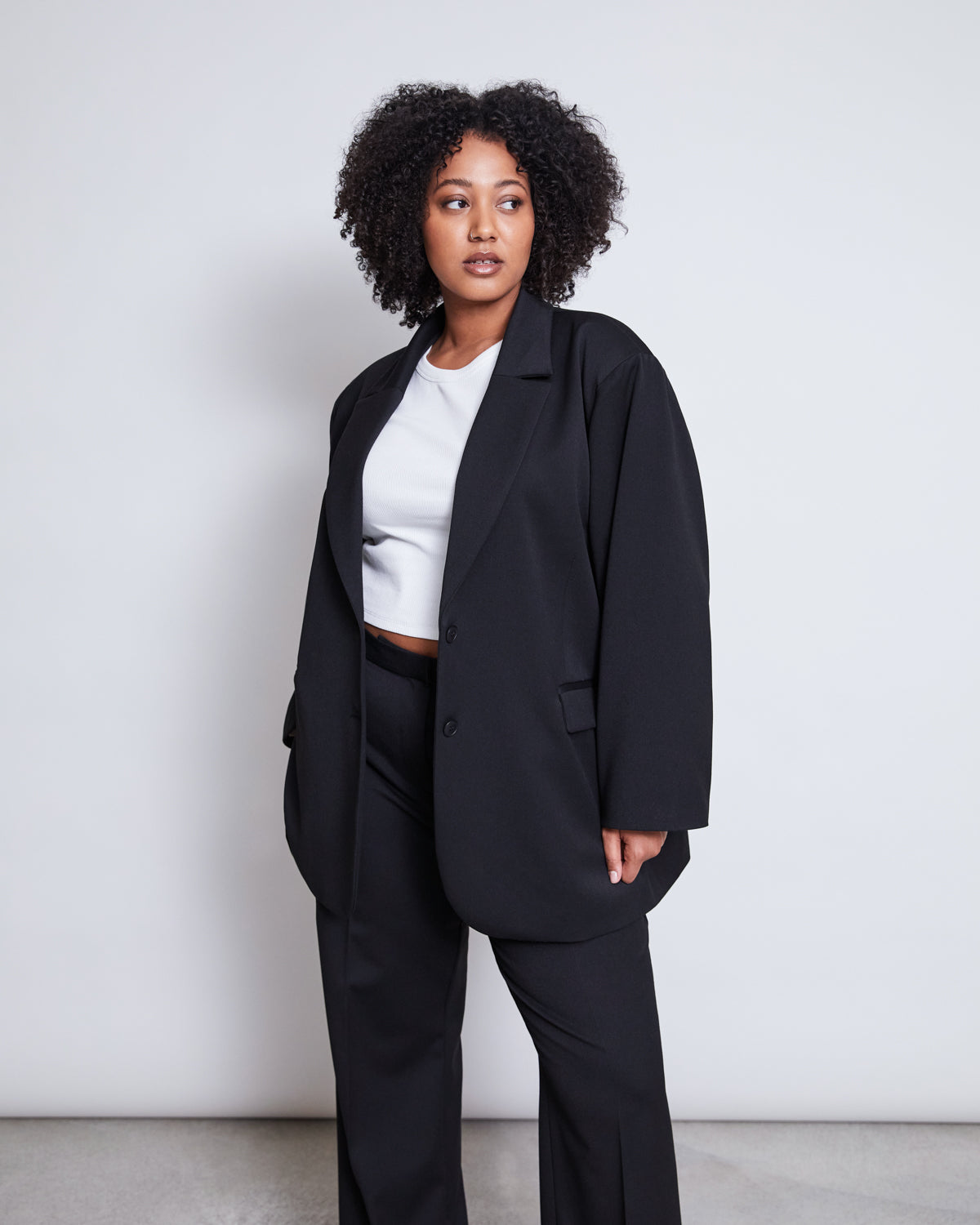 A woman with curly hair wears the jan-n-june BLAZER DURANCE BLACK, an oversized blazer made from recycled polyester, paired with matching pants and a white crop top. She stands against a plain light background, looking to the side.