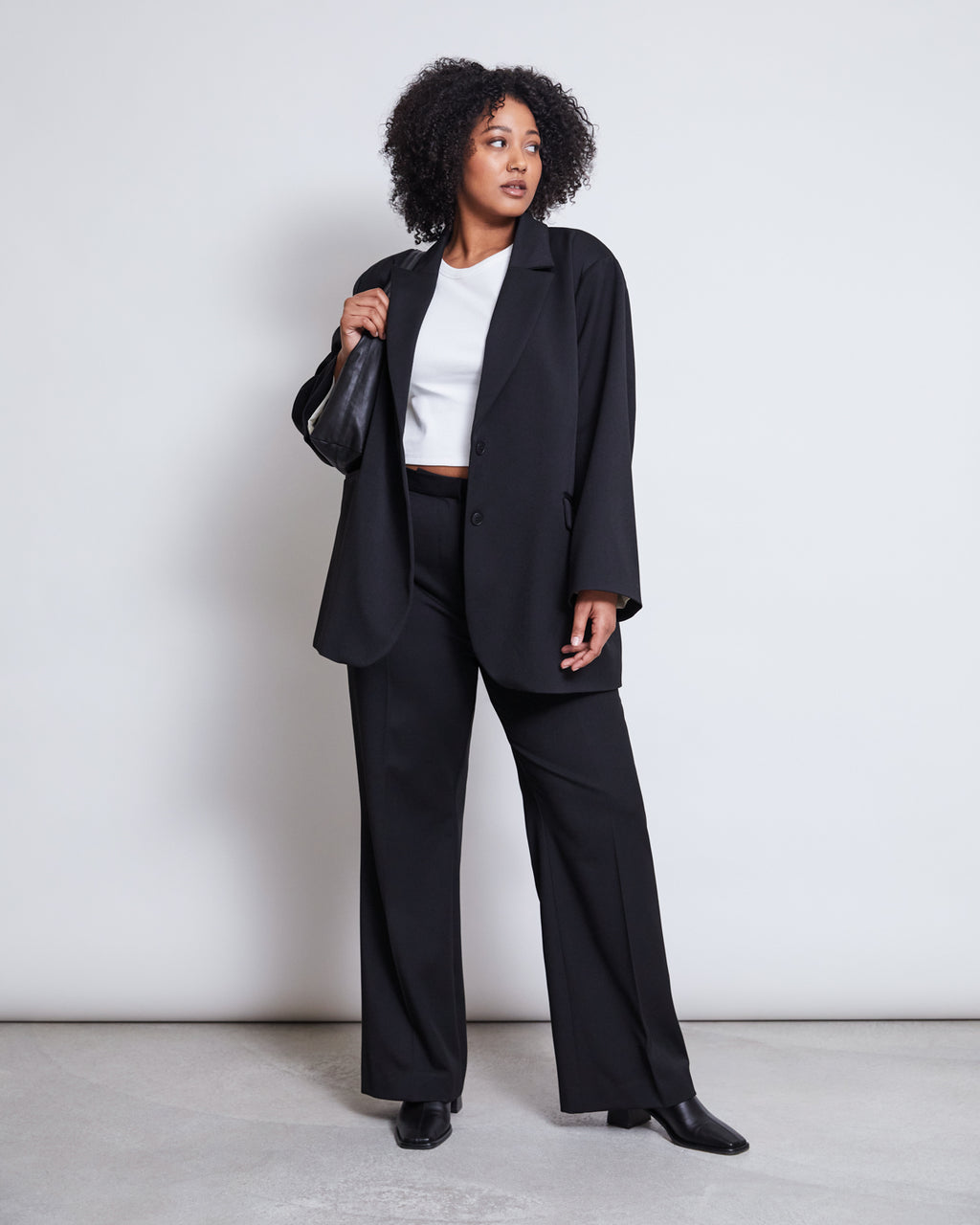 A woman with curly hair stands against a plain background, wearing the jan-n-june BLAZER DURANCE BLACK with matching trousers, a white crop top, black shoes, and holding a small black bag over her shoulder.
