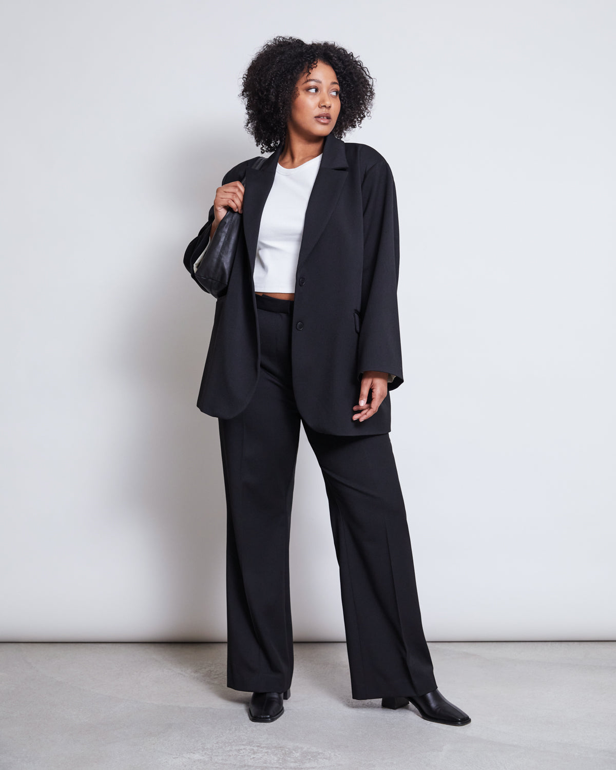 A woman with curly hair stands against a plain background, wearing the jan-n-june BLAZER DURANCE BLACK with matching trousers, a white crop top, black shoes, and holding a small black bag over her shoulder.