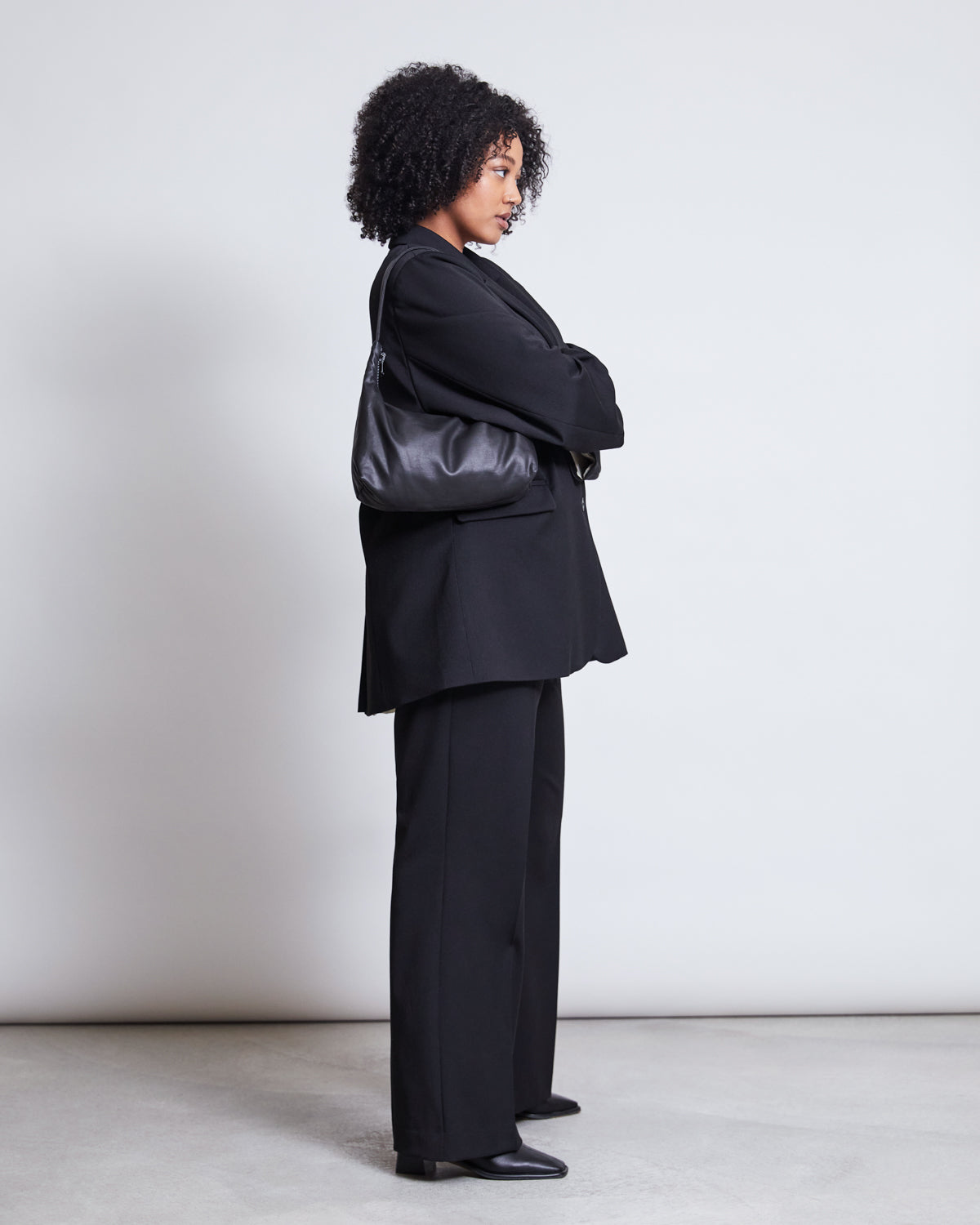 A woman with curly hair stands in profile against a plain background, wearing the jan-n-june BLAZER DURANCE BLACK over black trousers and boots. She carries a black shoulder bag and crosses her arms.