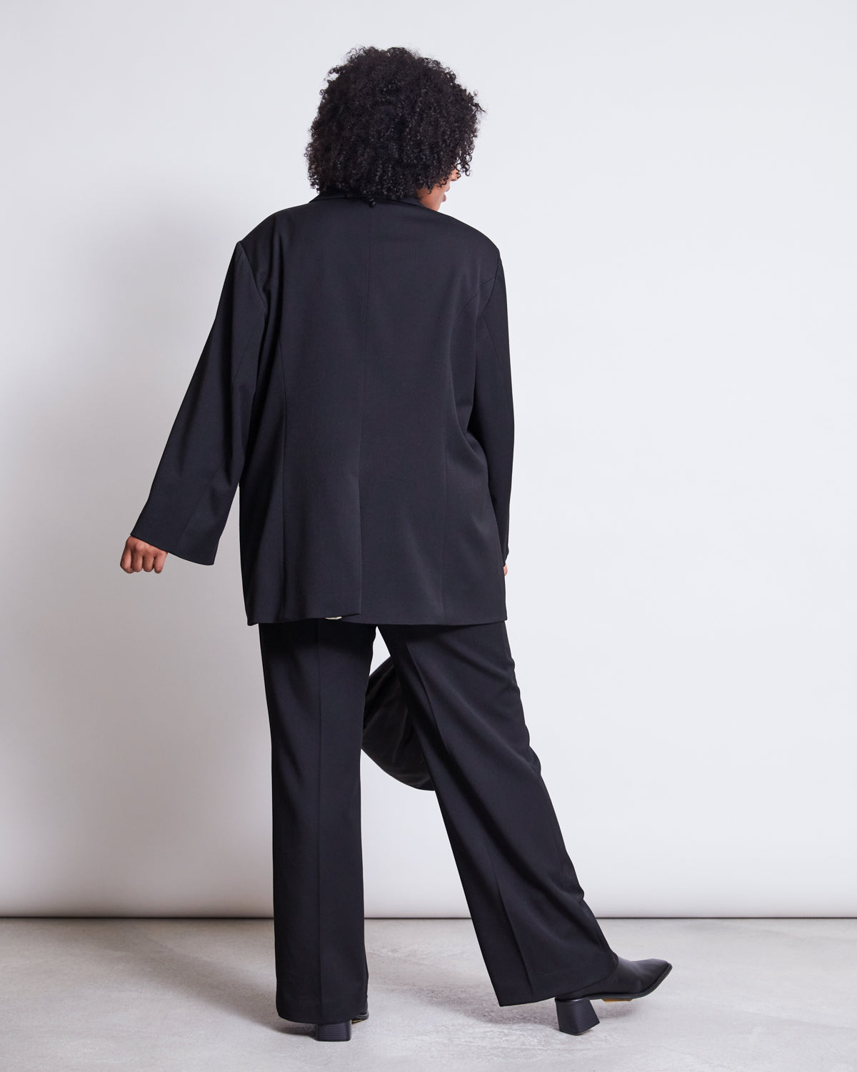 A person with curly hair wears the jan-n-june BLAZER DURANCE BLACK, an oversized blazer made from recycled polyester, paired with matching pants, standing against a plain white background with one foot slightly lifted.