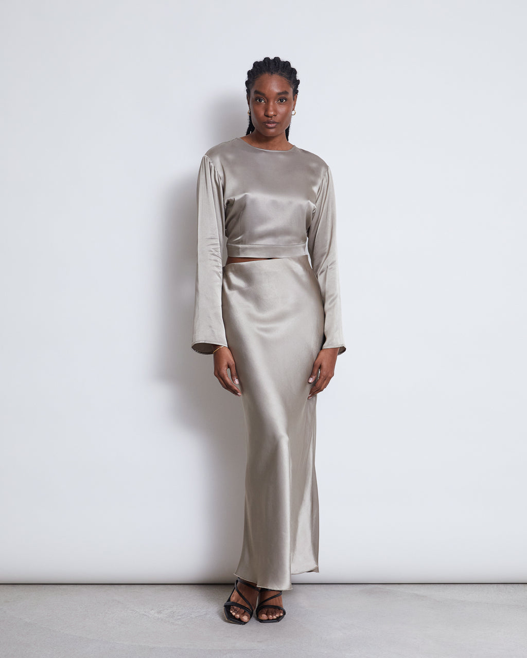 A woman stands against a white background wearing the jan-n-june OPEN BACK BLOUSE VIHM in silver grey with long sleeves and black strappy sandals, looking directly at the camera.