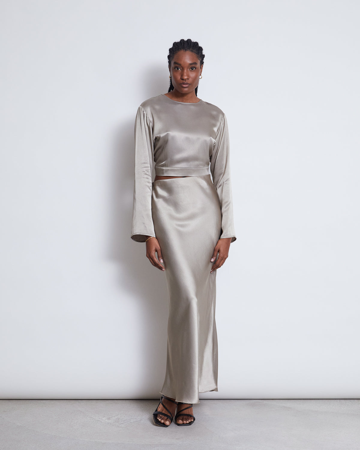 A woman stands against a white background wearing the jan-n-june OPEN BACK BLOUSE VIHM in silver grey with long sleeves and black strappy sandals, looking directly at the camera.