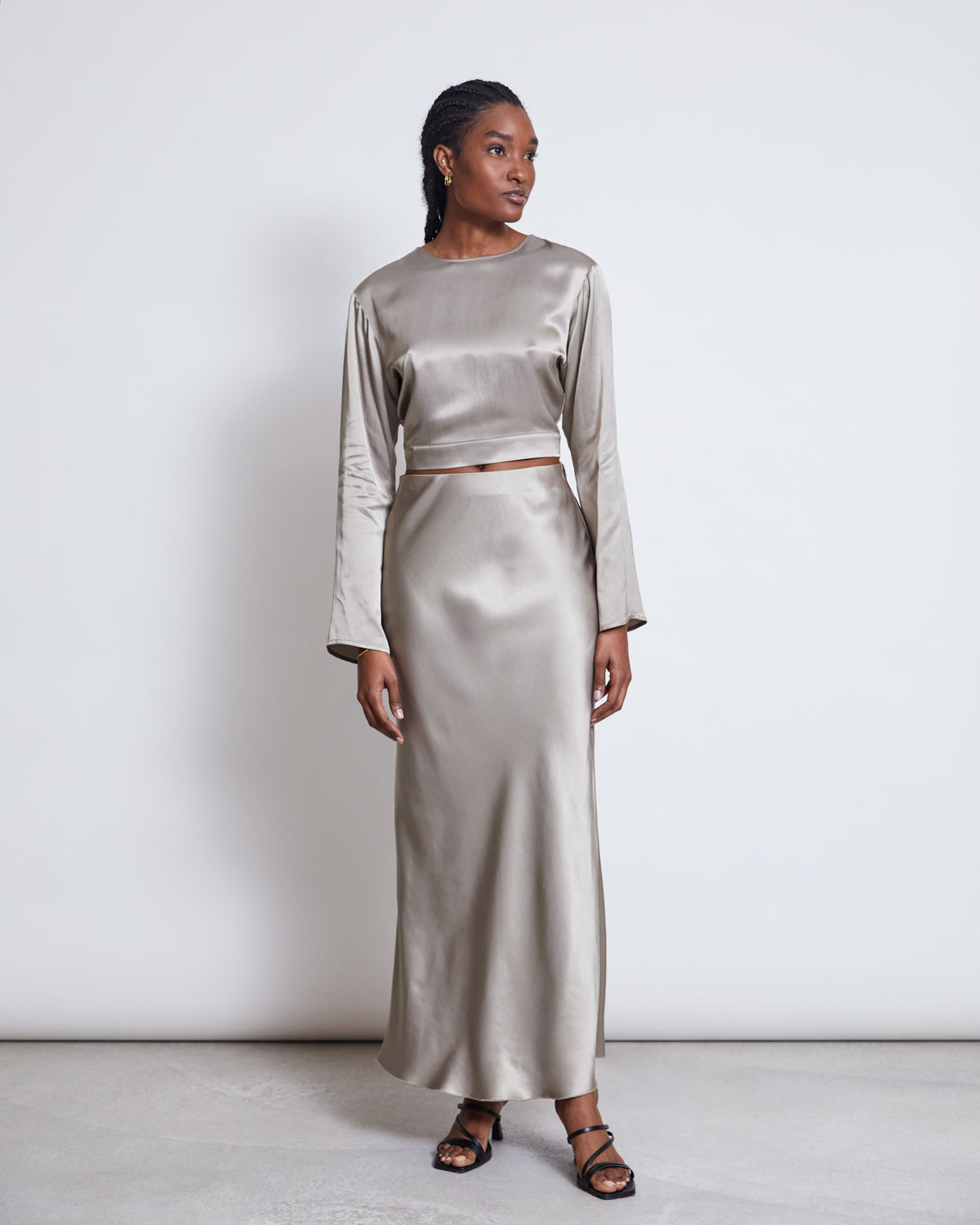 A woman stands against a plain light background wearing the jan-n-june OPEN BACK BLOUSE VIHM in silver grey with long sleeves, paired with black strappy sandals. She looks to her left with a relaxed expression.