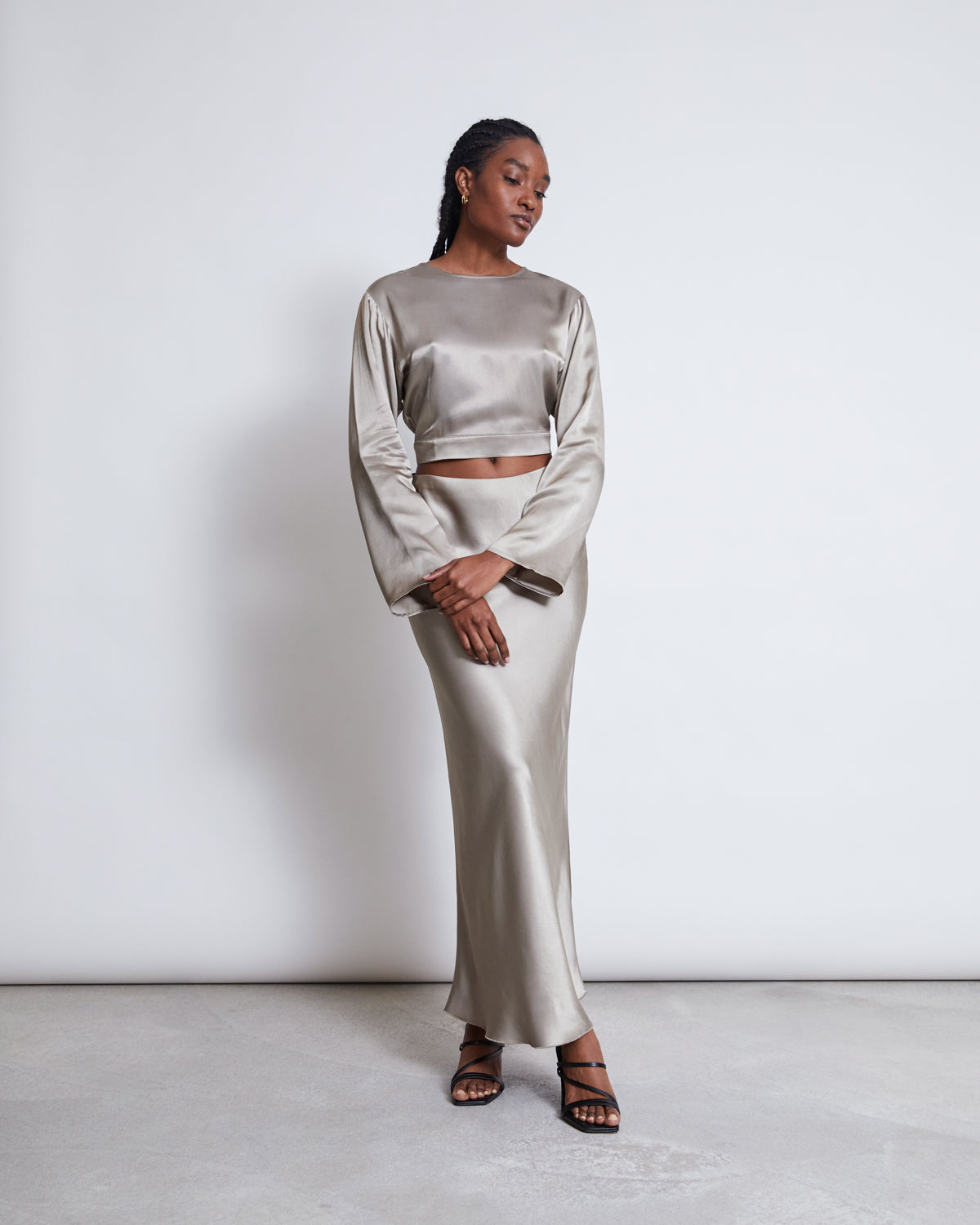 A woman wears jan-n-june’s OPEN BACK BLOUSE VIHM SILVER GREY—an ankle-length, long-sleeve sustainable blouse with a shiny silver finish made from Circulose®—paired with black strappy heels, standing against a white background.