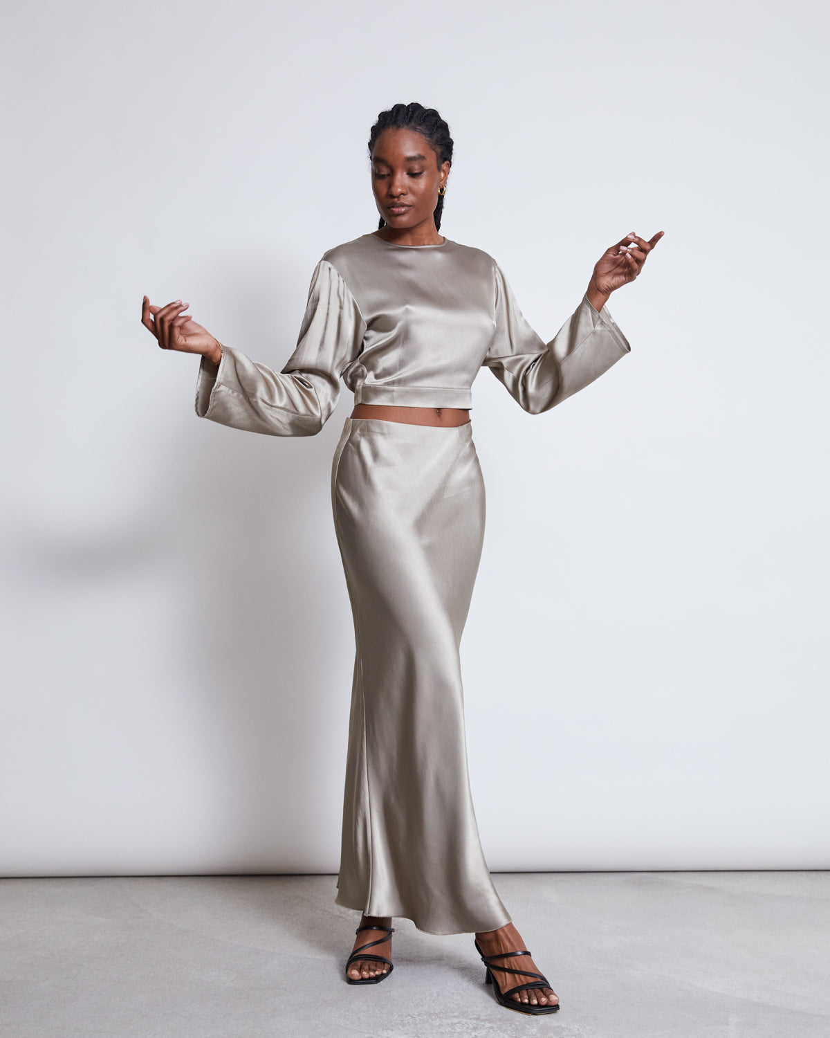 A woman poses gracefully in a long silver satin skirt and the jan-n-june OPEN BACK BLOUSE VIHM SILVER GREY, featuring flared sleeves, against a plain white background. She finishes the look with strappy black heels.