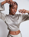 A woman in the jan-n-june OPEN BACK BLOUSE VIHM SILVER GREY poses gracefully with raised arms and spread fingers, set against a plain light background.