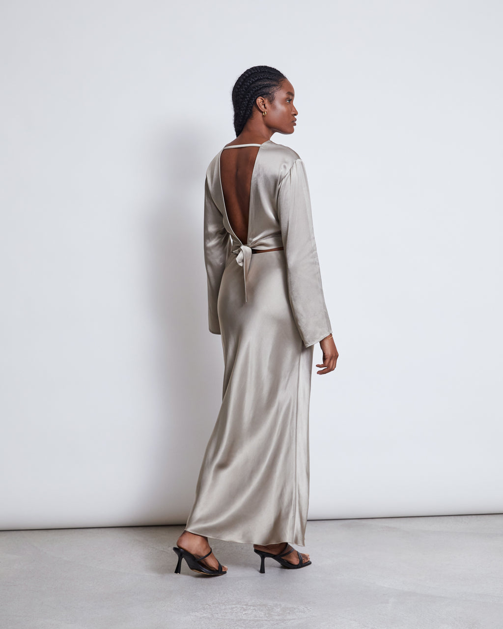 A woman in profile wears the jan-n-june OPEN BACK BLOUSE VIHM SILVER GREY, made of ENKA® Viscose. The long-sleeve, silver blouse features an open back tied at the waist. She has braided hair, black heeled sandals, and stands against a plain white background.