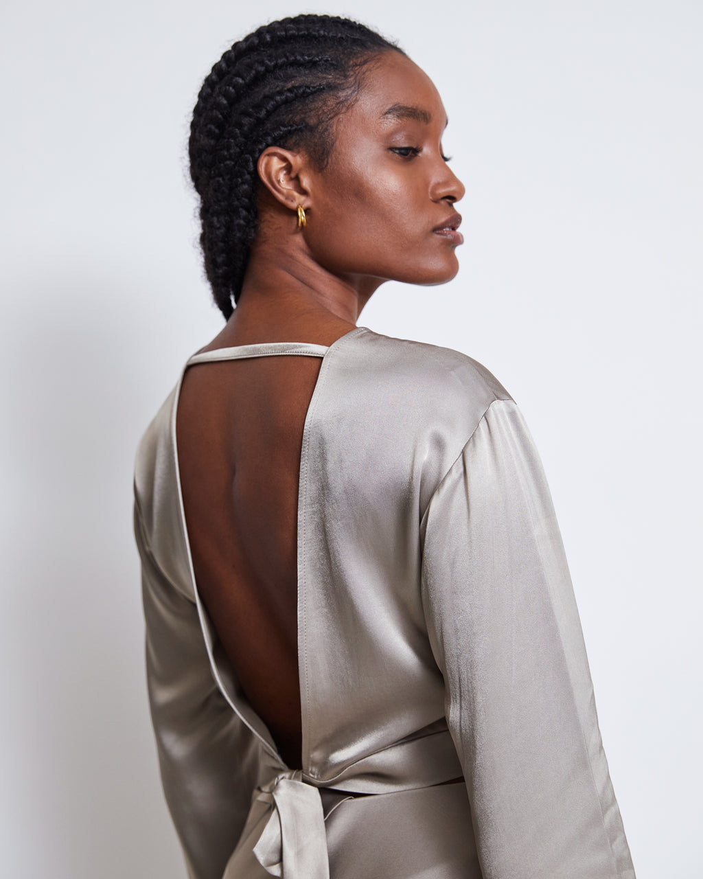 A woman with braided hair, gold hoop earrings, and a shiny silver grey jan-n-june OPEN BACK BLOUSE VIHM featuring a tie detail stands against a white background, looking over her shoulder.