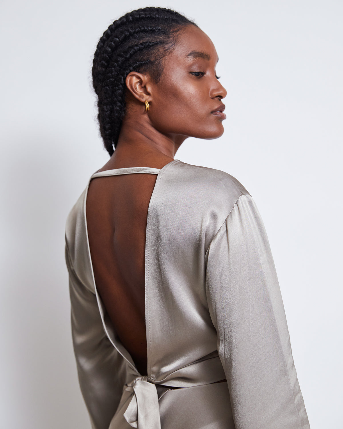 A woman with braided hair, gold hoop earrings, and a shiny silver grey jan-n-june OPEN BACK BLOUSE VIHM featuring a tie detail stands against a white background, looking over her shoulder.