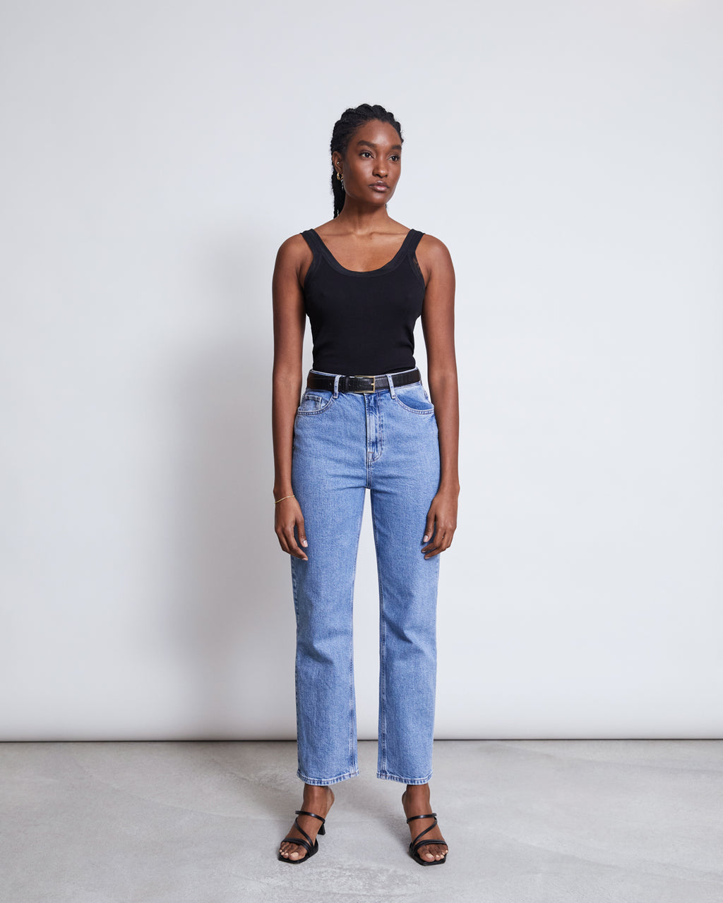 A woman stands against a plain light gray background, wearing the jan-n-june MESH DETAIL TOP LLUVIA BLACK with high-waisted blue jeans, a black belt, and black heeled sandals. She has a neutral expression and gazes slightly to the side.