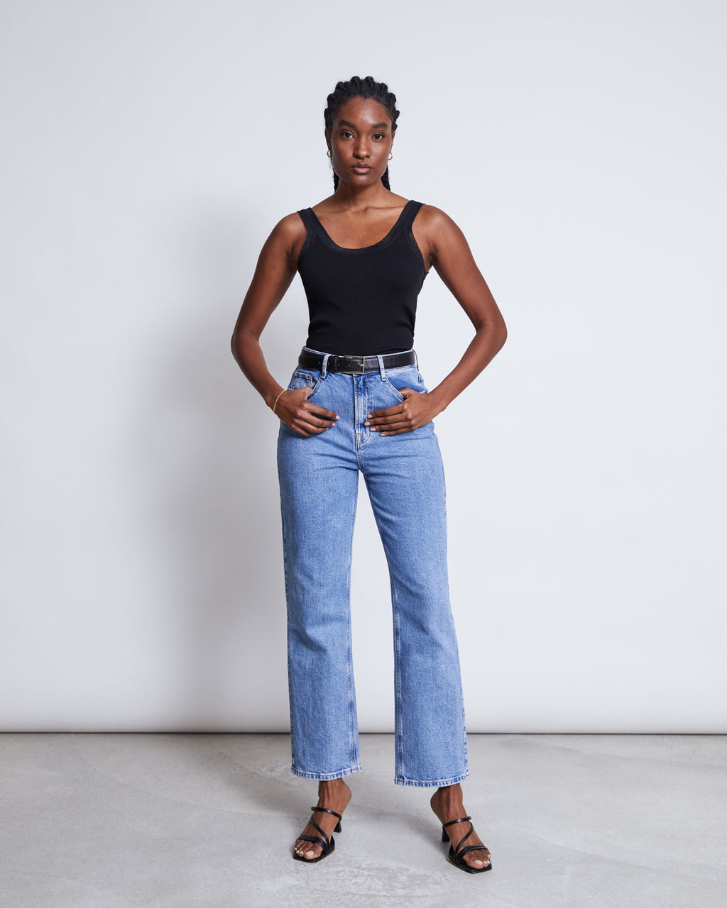 A woman poses confidently against a plain background, wearing the jan-n-june MESH DETAIL TOP LLUVIA BLACK, light blue high-waisted jeans, a black belt, and black heeled sandals. Her hands rest on her hips as she faces the camera.