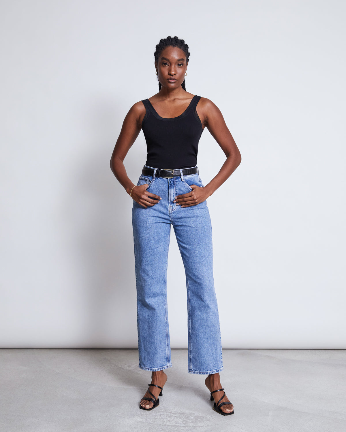 A woman poses confidently against a plain background, wearing the jan-n-june MESH DETAIL TOP LLUVIA BLACK, light blue high-waisted jeans, a black belt, and black heeled sandals. Her hands rest on her hips as she faces the camera.