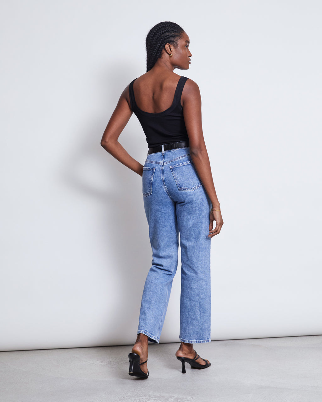 A woman with braided hair faces away from the camera indoors, wearing the jan-n-june MESH DETAIL TOP LLUVIA BLACK, blue high-waisted jeans, and black heeled sandals against a plain light-colored wall.