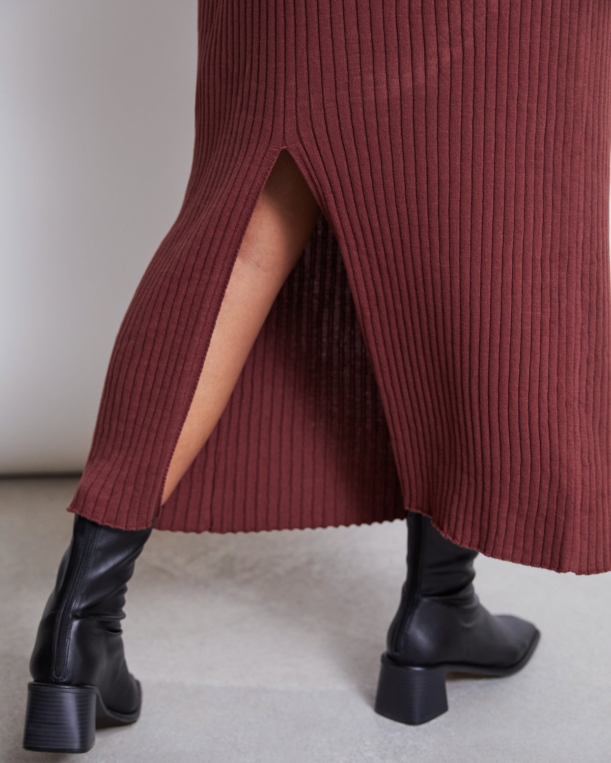 A person wearing the jan-n-june MIDI KNIT SKIRT EBBA INTENSE RUST GOTS with a back slit and black heeled ankle boots, standing on a light-colored floor.