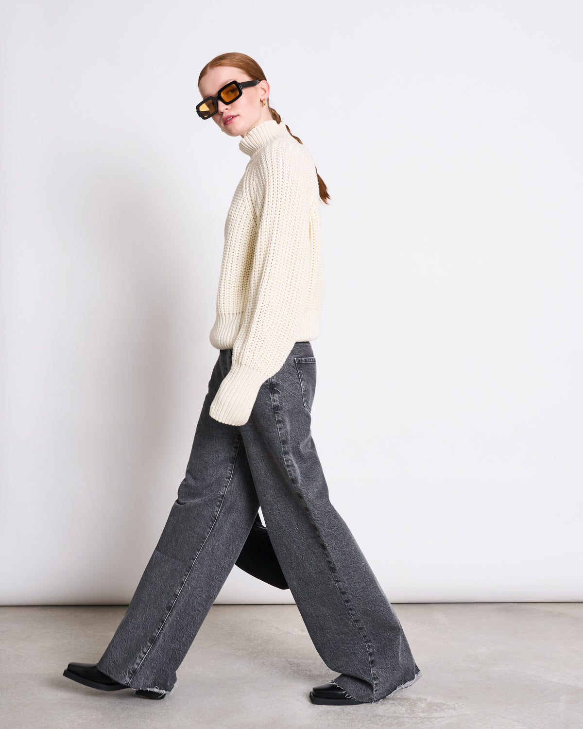 A person with long hair in a ponytail wears large sunglasses, gray wide-leg jeans, and the jan-n-june JUMPER OLA OFFWHITE GOTS chunky turtleneck made from organic cotton, walking against a plain white background.