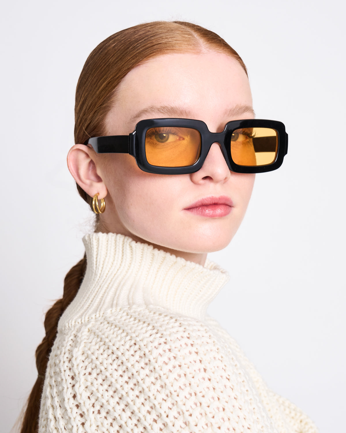 A woman with red hair in a low ponytail wears chunky black square sunglasses with yellow lenses, gold hoop earrings, and the jan-n-june JUMPER OLA OFFWHITE GOTS turtleneck jumper against a plain background.