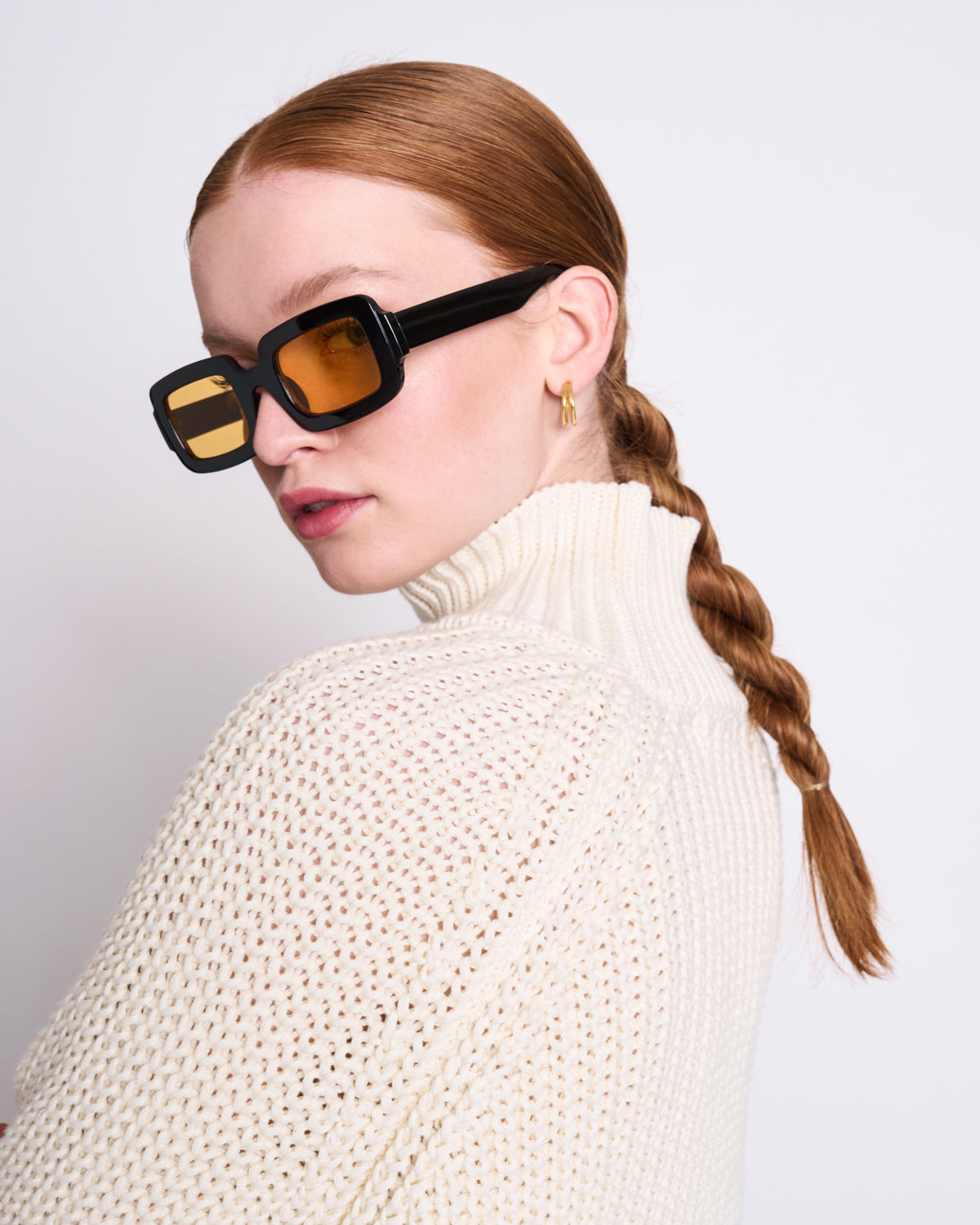 A woman with red hair in a braid wears oversized black sunglasses and the jan-n-june JUMPER OLA OFFWHITE GOTS turtleneck, looking over her shoulder against a plain white background.