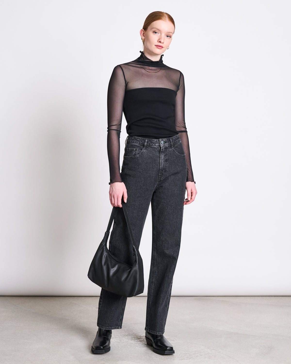 A person with red hair wears the jan-n-june MESH TURTLENECK PLUIE BLACK over a black tank, paired with high-waisted black jeans, black shoes, and a black handbag, standing against a plain white background.