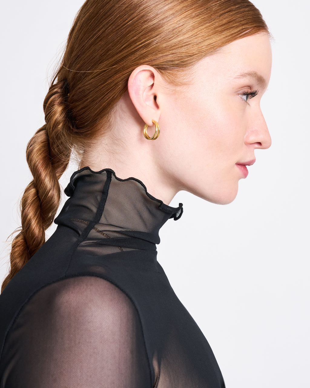 A woman with fair skin and red hair in a twisted ponytail is shown in profile, wearing the jan-n-june MESH TURTLENECK PLUIE BLACK with ruffled trim and gold hoop earrings against a plain white background.
