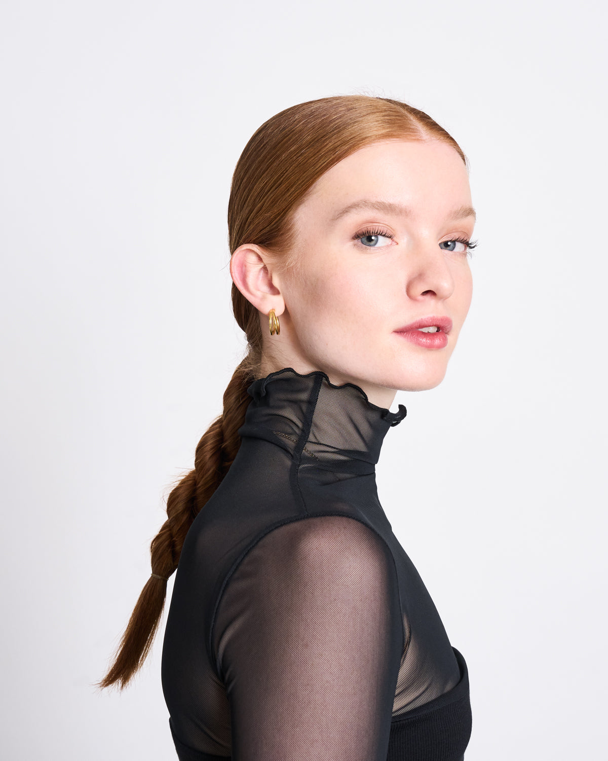 A young woman with fair skin and red braided hair wears the jan-n-june MESH TURTLENECK PLUIE BLACK, made from recycled polyester, plus gold hoop earrings, looking over her shoulder against a white background.