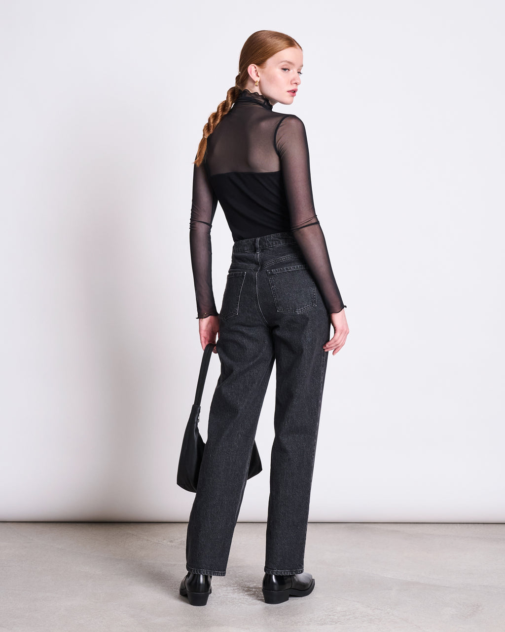 A woman with braided hair stands facing away, wearing the jan-n-june MESH TURTLENECK PLUIE BLACK, paired with high-waisted dark jeans, black shoes, and a black handbag, against a plain white background.
