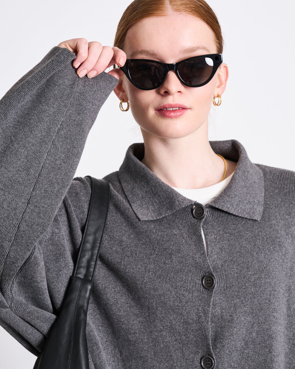 A red-haired woman with her hair tied back wears black cat-eye sunglasses, gold hoop earrings, and the jan-n-june CARDIGAN ANJO DARK GREY GOTS—an oversized gray organic cotton knit jacket—while carrying a black shoulder bag.