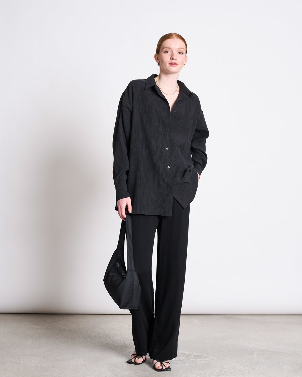 A woman with red hair stands against a plain backdrop wearing the jan-n-june CRINKLE SHIRT SKUR BLACK, wide-leg black pants, black sandals, carrying a black bag with one hand in her pocket.