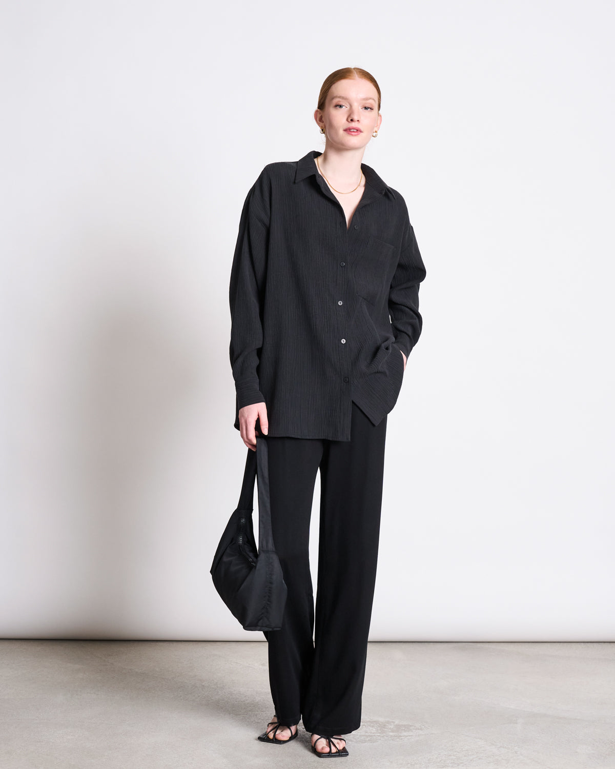 A woman with red hair stands against a plain backdrop wearing the jan-n-june CRINKLE SHIRT SKUR BLACK, wide-leg black pants, black sandals, carrying a black bag with one hand in her pocket.