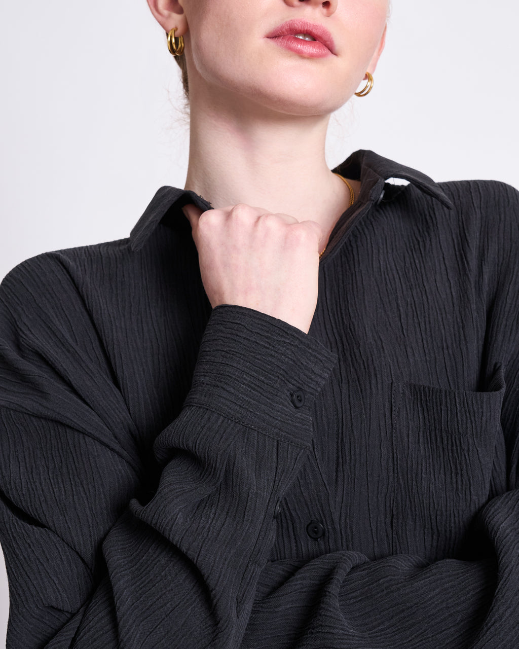 A person poses against a plain light background, wearing the jan-n-june CRINKLE SHIRT SKUR BLACK and gold hoop earrings, with one hand lightly gripping their collar.