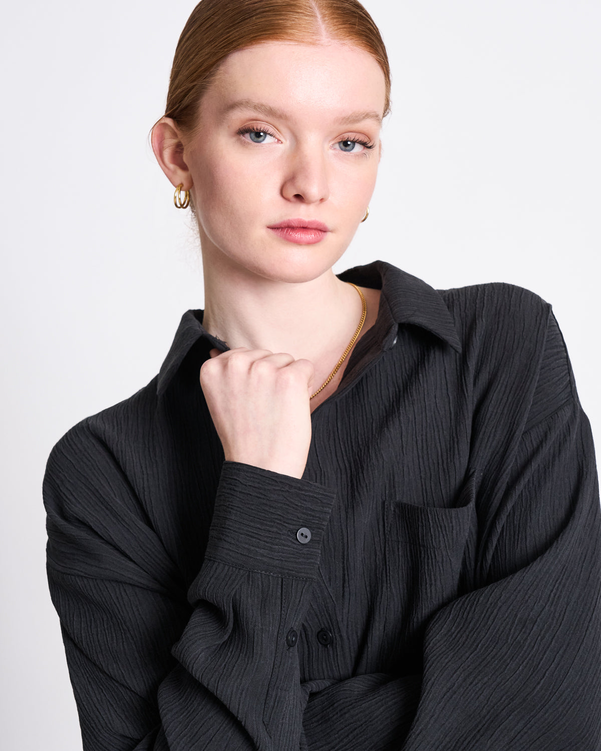 A person with light skin and red hair tied back wears gold earrings and the jan-n-june CRINKLE SHIRT SKUR BLACK, made from soft Tencel™ Lyocell fabric. They look at the camera neutrally, resting their fist under their chin.