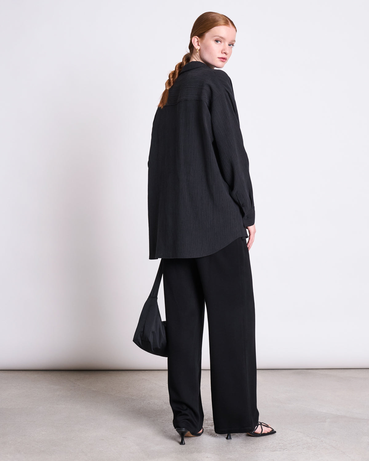 A person with long, braided hair wears the jan-n-june CRINKLE SHIRT SKUR BLACK, loose black pants, strappy heels, and carries a black bag. They stand on a light floor, facing away while looking back over their shoulder.