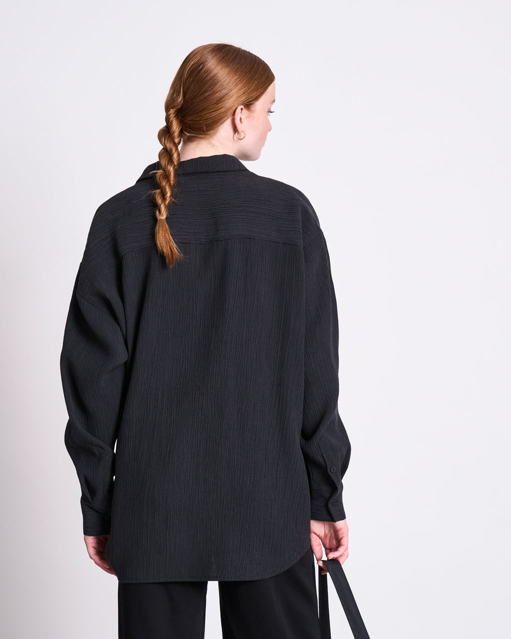 A woman with long red hair in a braid stands with her back to the camera, wearing the jan-n-june CRINKLE SHIRT SKUR BLACK and black pants against a plain white background.