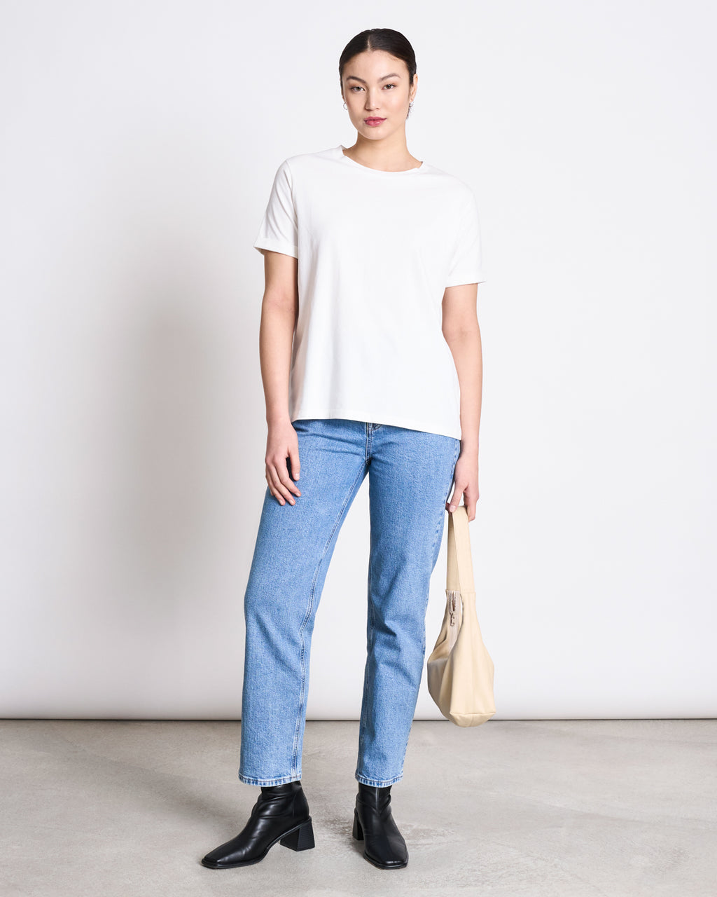 A person stands against a plain white wall, wearing the jan-n-june BRUSHED T-SHIRT BOY PLAIN WHITE GOTS, blue jeans, black ankle boots, and holding a beige handbag with their hair tied back.